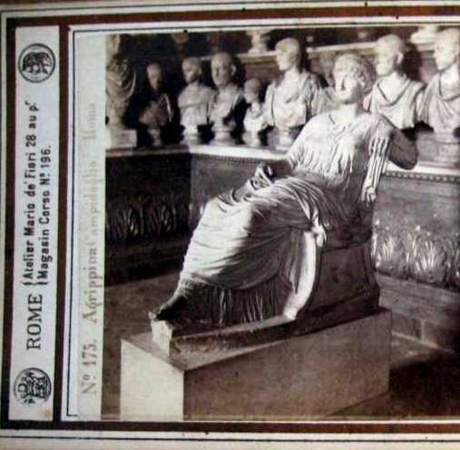 Old photograph of a seated marble Roman lady in long flowing drapery, her head bent and her elbow propped on her knee, in a room with rows of portrait busts behind her