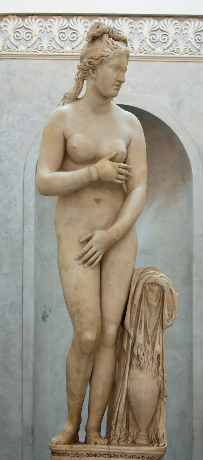 Marble statue of a nude Venus covering her breast and groin with her hands, hair gathered in a knot with a long lock over one shoulder, an urn draped with cloth at her side