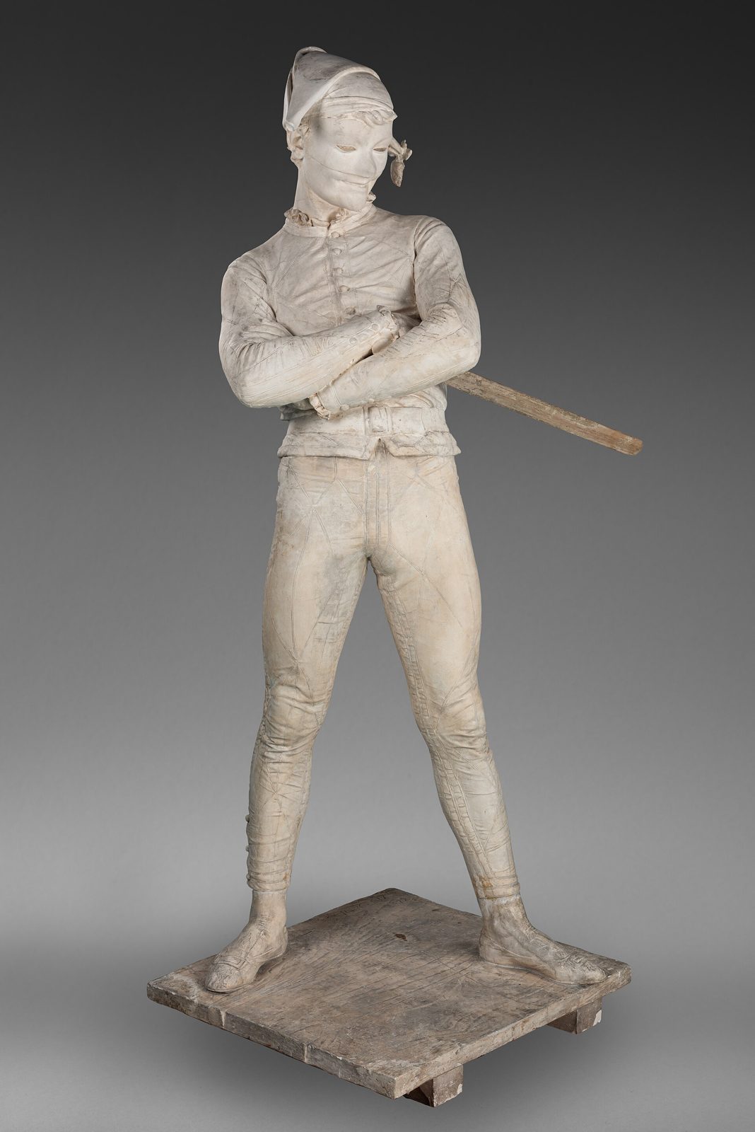 A standing white plaster Harlequin in mask, tight suit and soft cap, legs apart, arms crossed over his chest, a wooden bat in his right hand, head tilted mockingly to one side, on a rough plank base
