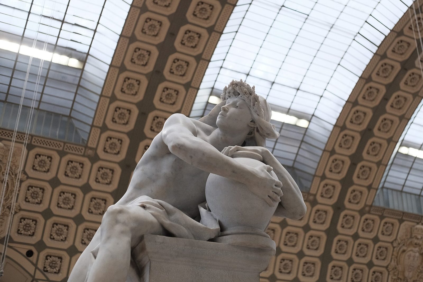 A seated marble winged youth, nude, crouched on a rocky base with a finger raised to his lips in a gesture of silence, his head bowed over the tomb he guards