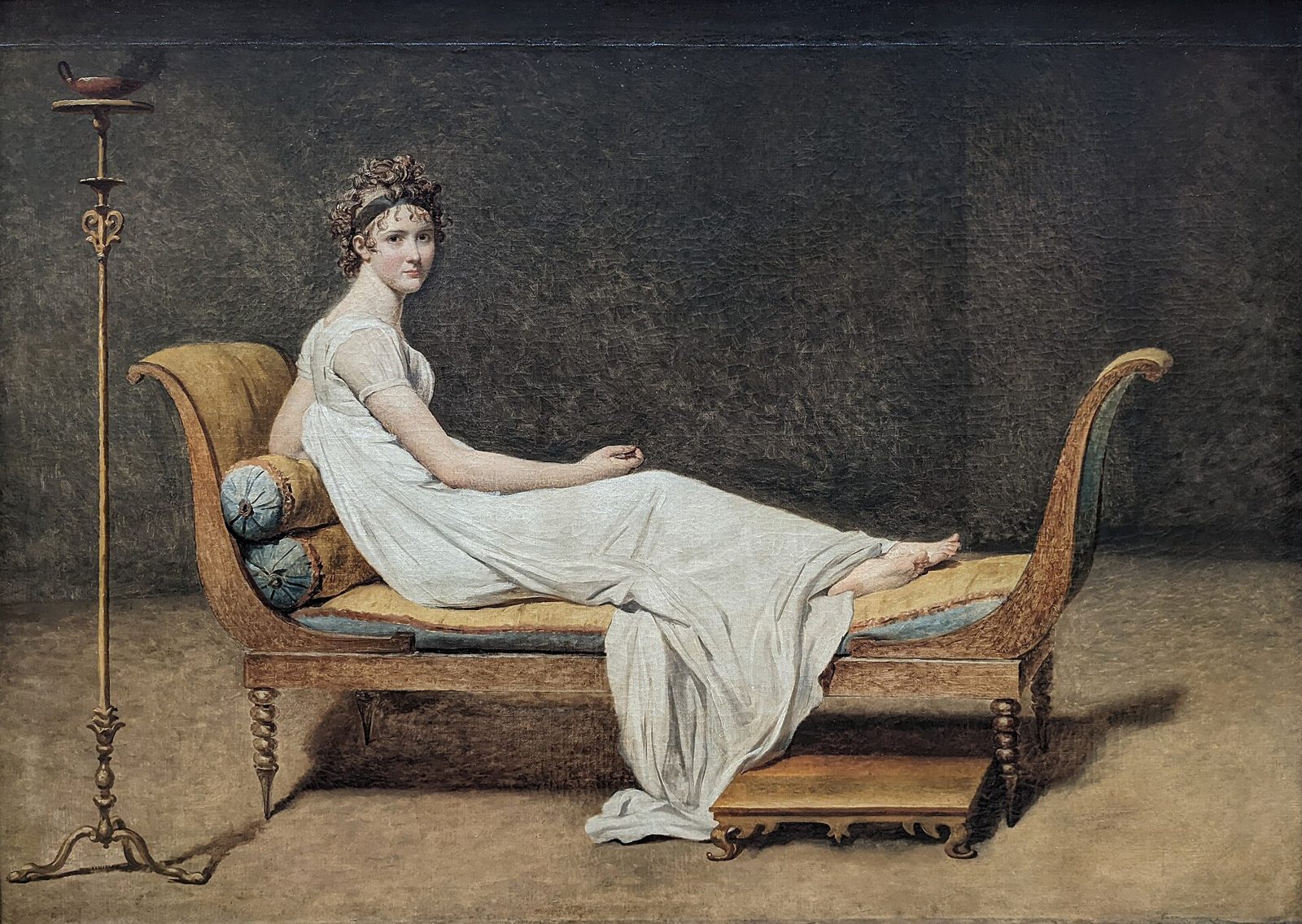 A young woman with her hair bound in a classical knot reclines on a bare Empire chaise longue in a plain white sleeveless dress, bare feet, looking toward the viewer, against an empty grey-green wall